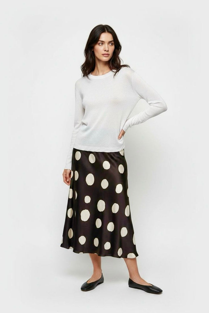 A woman modeling a white long-sleeve knit top and a black midi skirt with large cream polka dots, paired with black flats, against a plain light gray studio background.