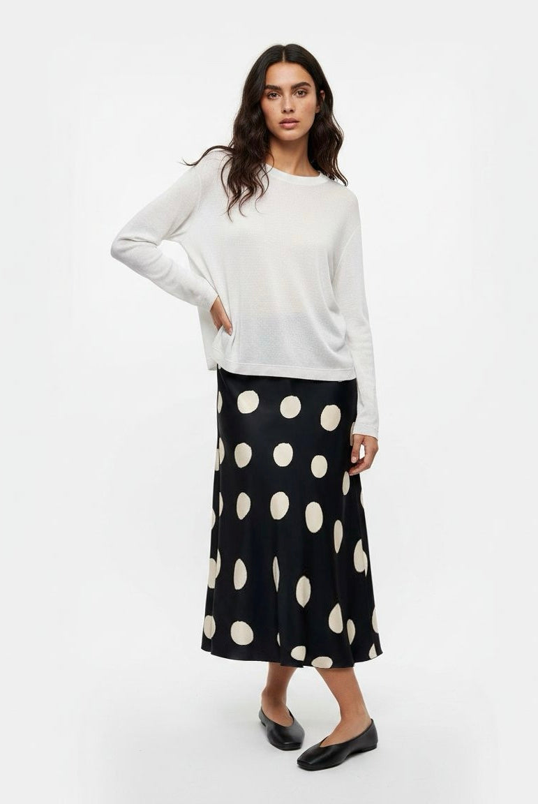 A woman modeling a white long-sleeve knit top and a black midi skirt with large cream polka dots, paired with black flats, against a plain white studio background.