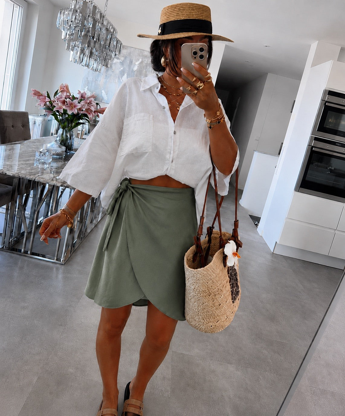 A woman wearing a straw fedora hat, a white cropped button-up shirt, an olive green wrap mini skirt
