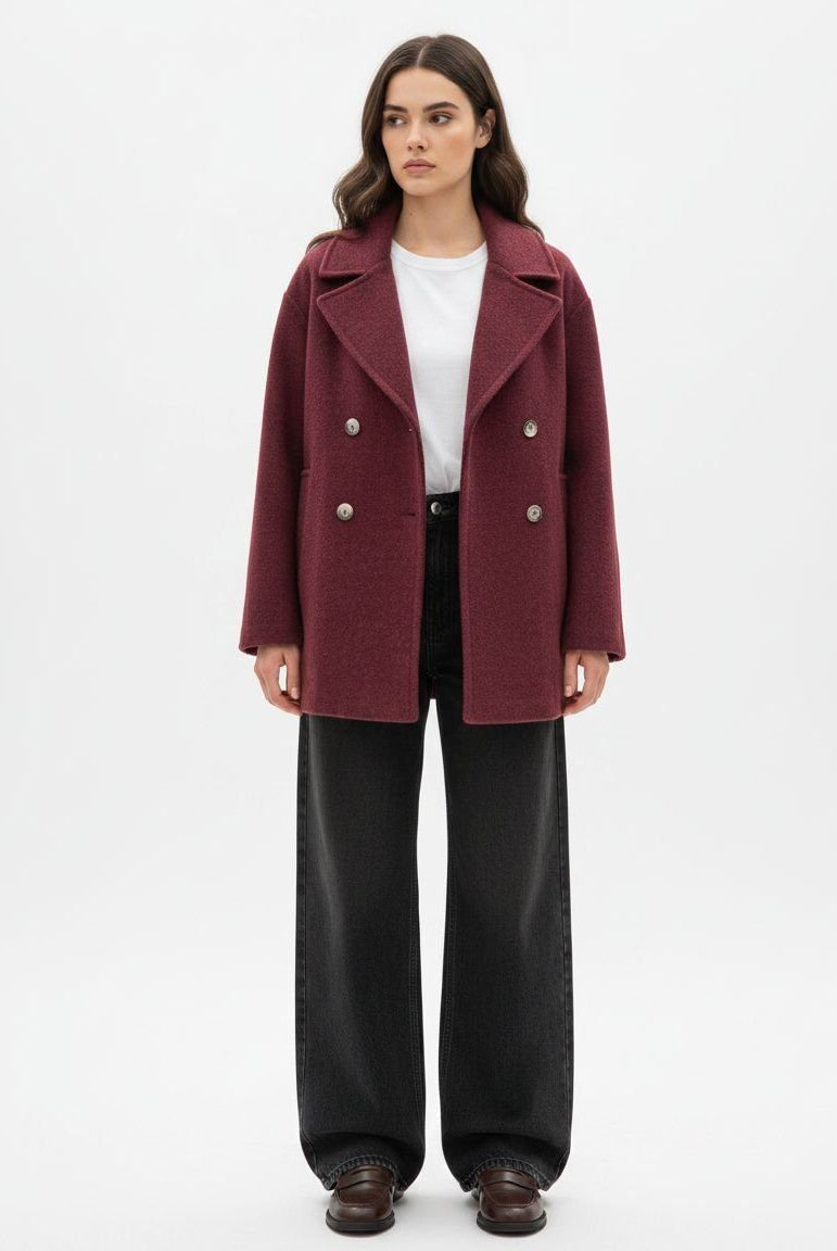 A woman stands wearing a burgundy double-breasted coat over a white top and black wide-leg trousers.