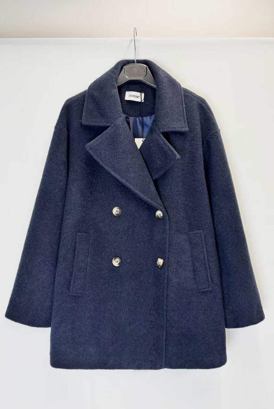 A navy blue double-breasted coat with silver buttons, hanging on a black hanger against a plain white wall.