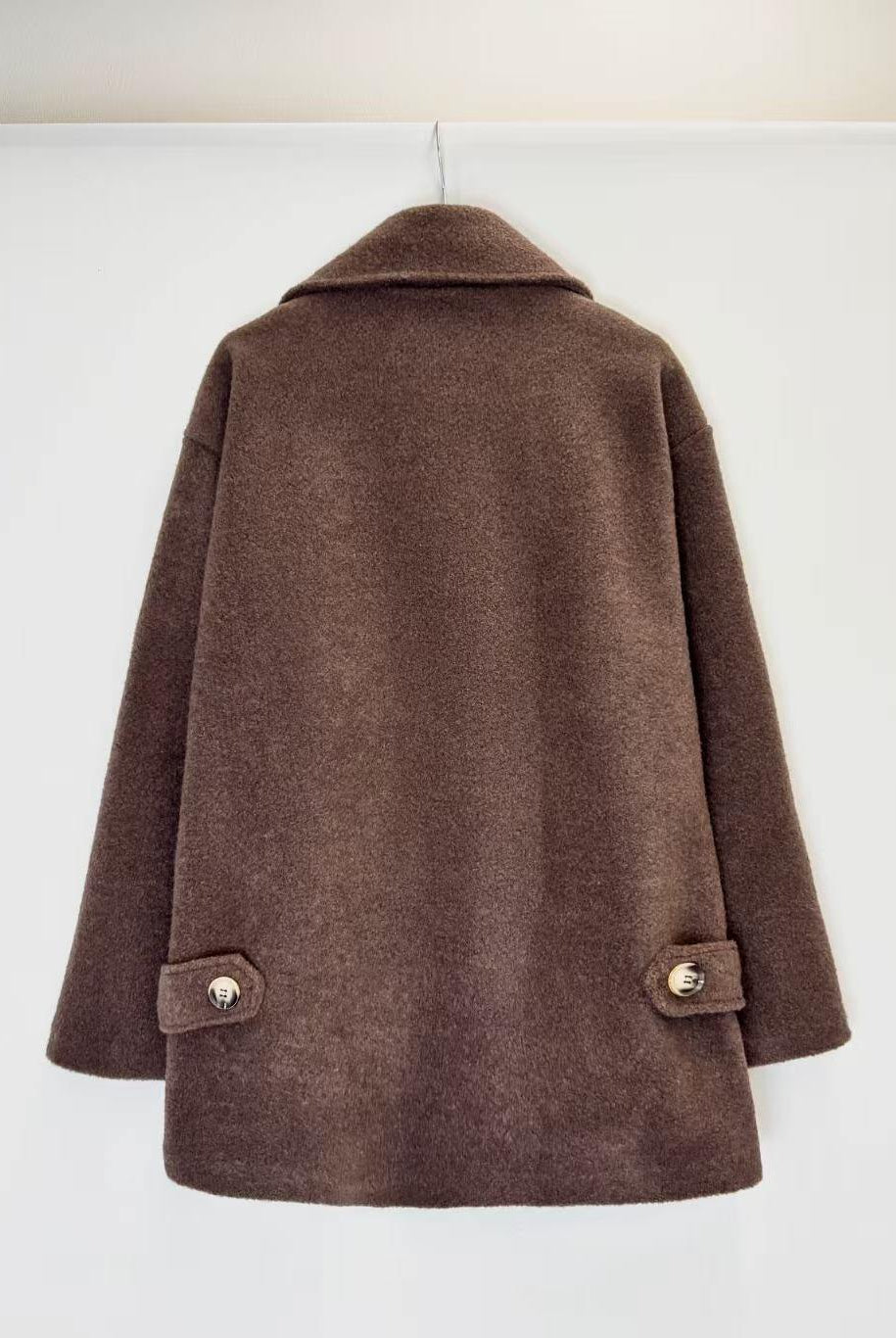 A brown wool-blend coat with a wide collar and buttoned flap pockets on the sleeves, hanging against a plain white wall.