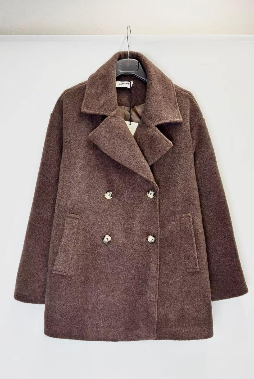 A brown double-breasted coat with wide lapels and buttons, hanging on a black hanger against a plain white wall.