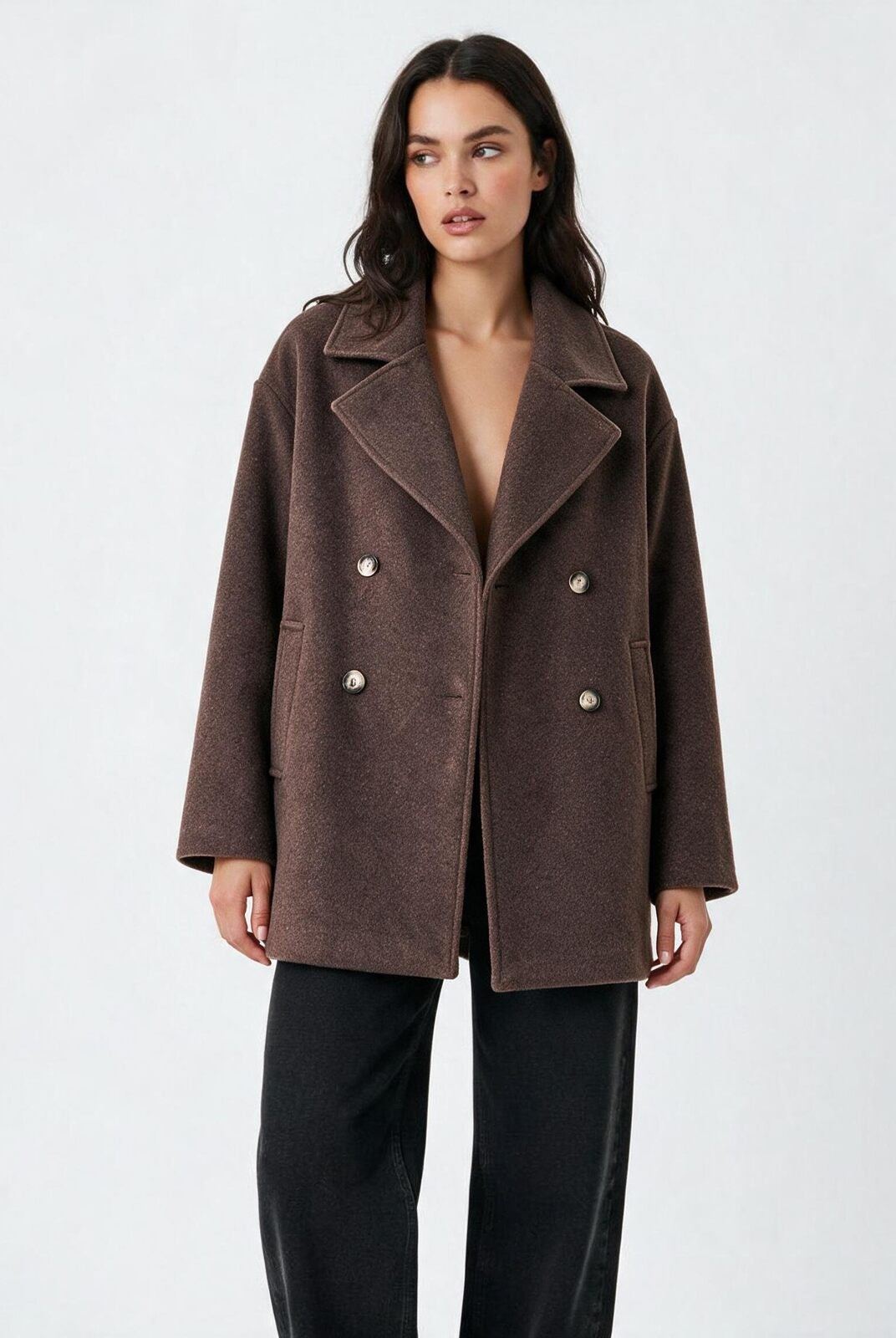 A woman modeling a brown double-breasted coat with silver buttons, paired with black trousers, against a plain light gray studio background.