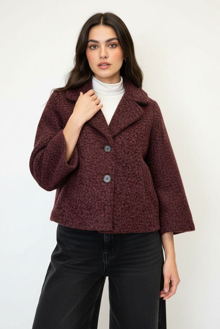 A woman modeling a textured, cropped dark burgundy jacket with wide lapels and three front buttons, worn over a white turtleneck and black wide-leg jeans, against a plain white background.