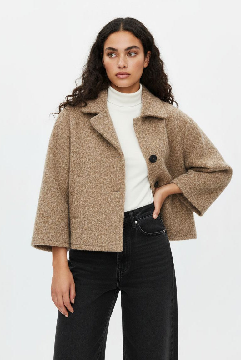 A woman modeling a cropped, textured beige coat with wide three-quarter sleeves, worn over a white turtleneck and black jeans, against a plain white studio background.