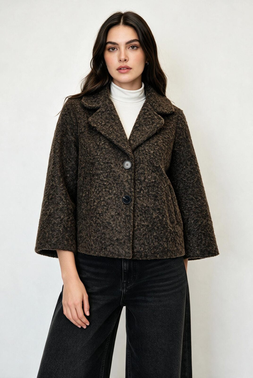A woman modeling a cropped, dark brown textured tweed jacket with wide three-quarter sleeves, worn over a white turtleneck and black wide-leg jeans, against a plain light gray background.