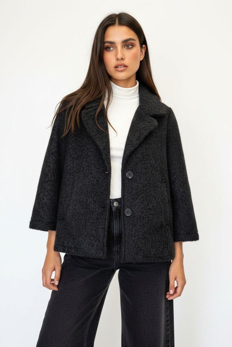 A woman modeling a dark charcoal gray textured wool-blend coat with a white turtleneck and black wide-leg jeans against a plain white studio background.