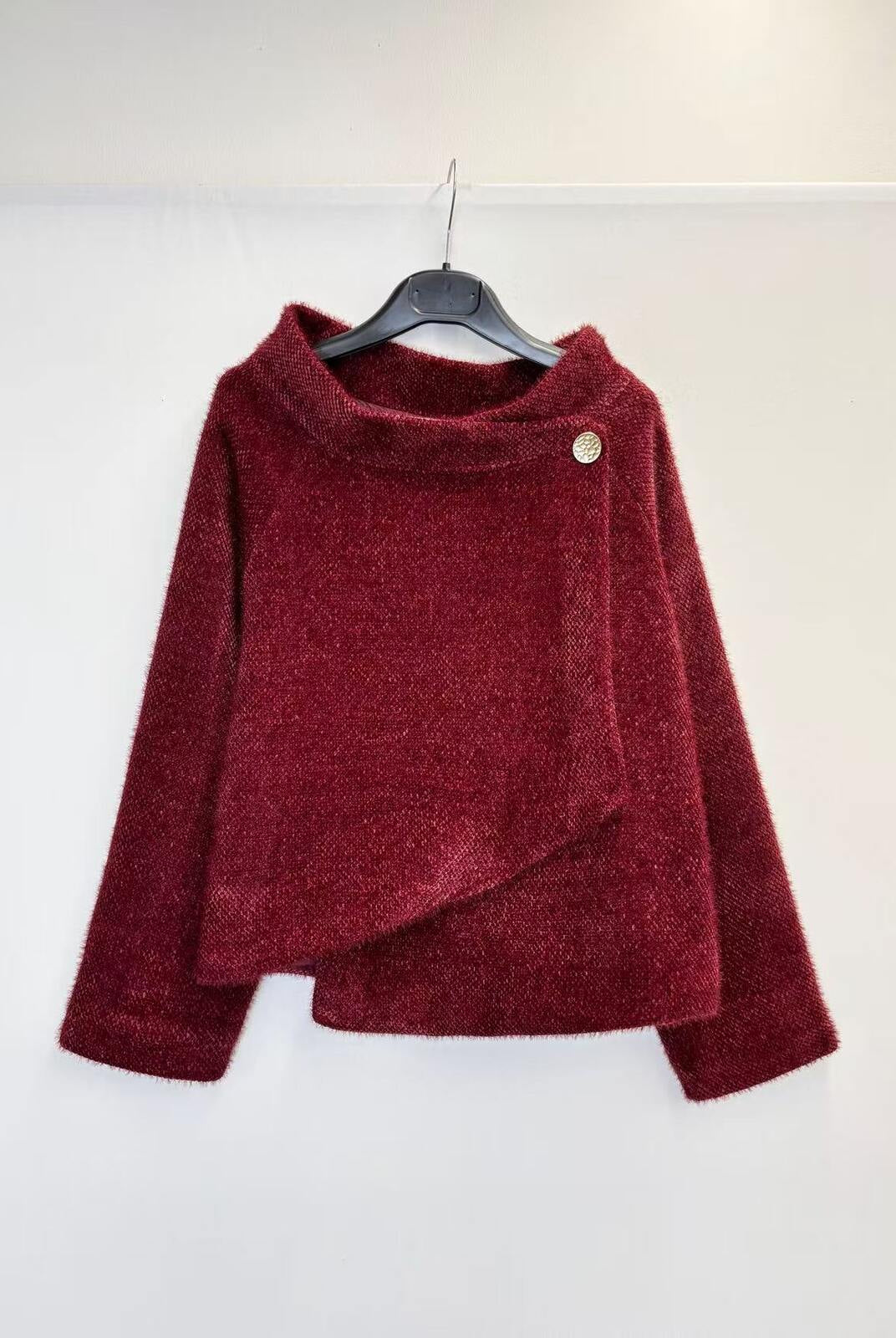 A textured, deep burgundy long-sleeve sweater with an asymmetrical wrap-style hem and a single decorative button, hanging on a black hanger against a plain white wall.