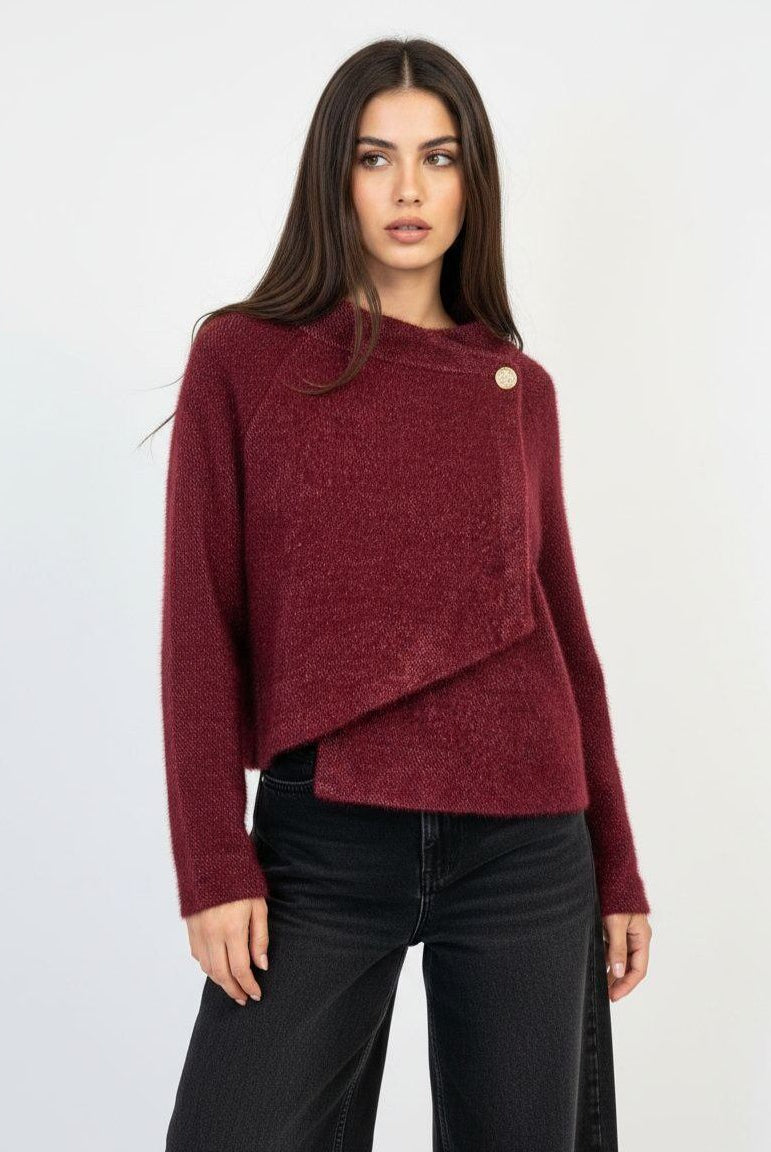 A woman modeling a textured, off-the-shoulder burgundy knit sweater with a single gold button detail, paired with black jeans, against a plain white studio background.