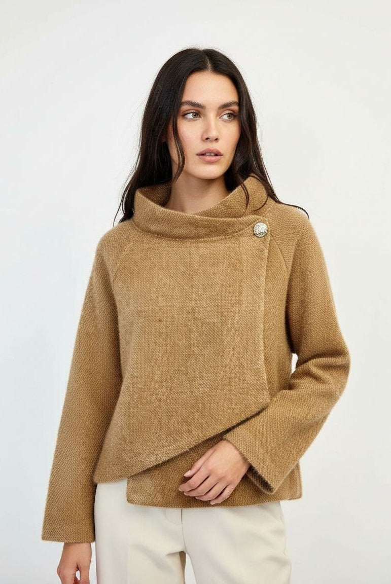 A woman wearing a camel-colored knit sweater with a cowl neck and asymmetrical button detail, paired with cream pants, against a plain light gray background.