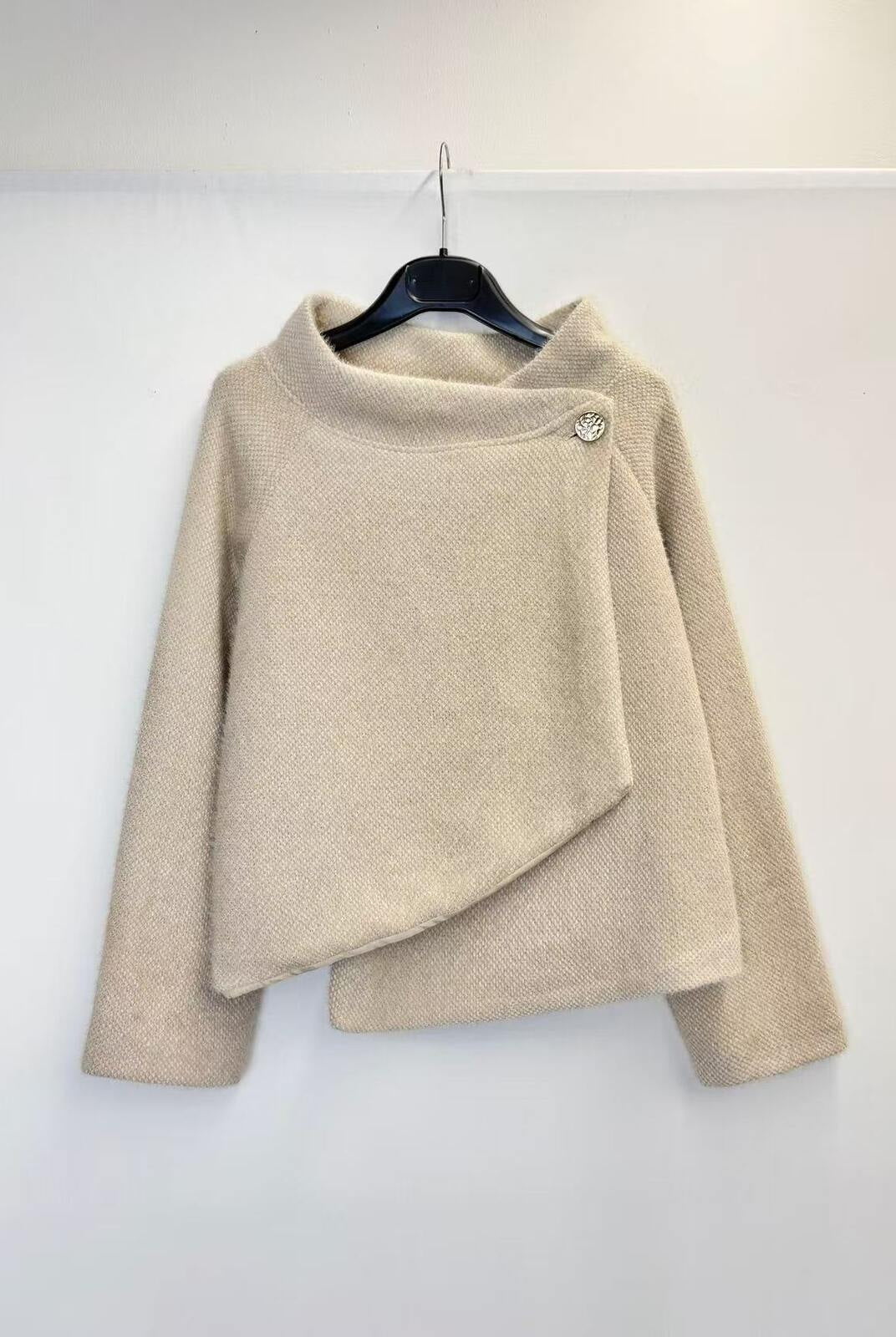 A beige textured knit sweater with a high mock neck and asymmetrical draped front detail, hanging on a black hanger against a plain white wall.