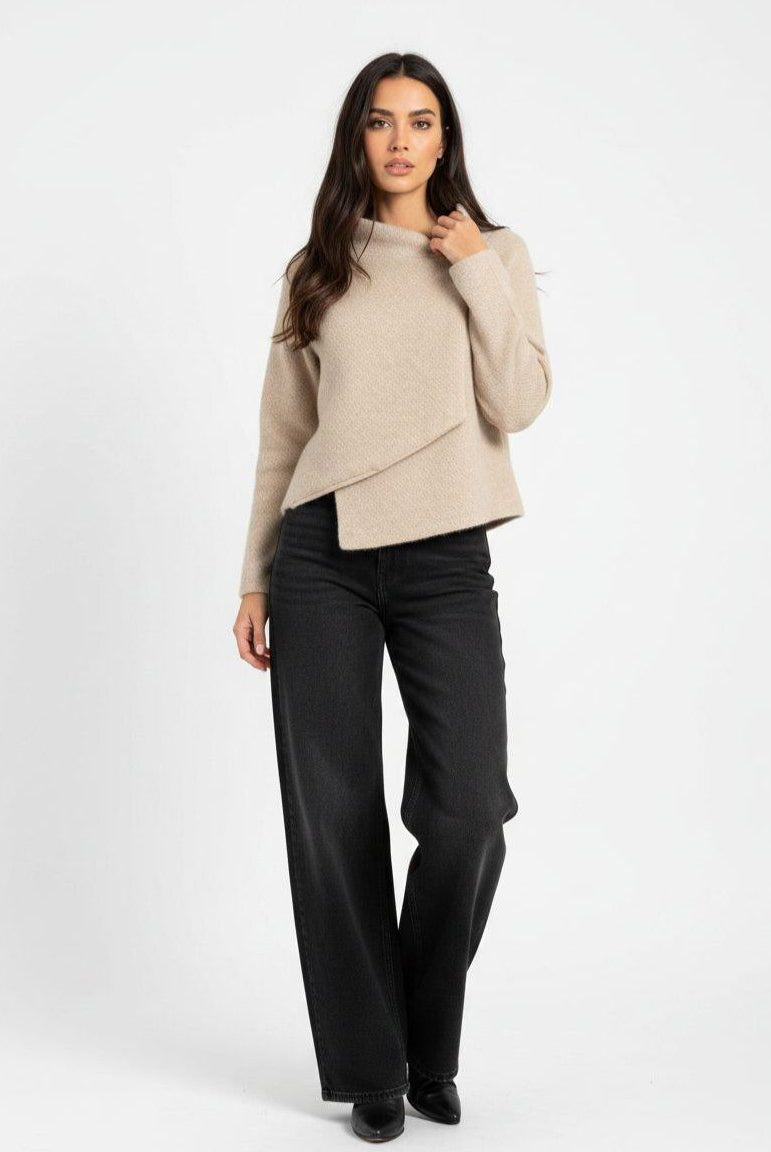 A woman modeling a light beige asymmetrical wrap-front knit sweater with black wide-leg trousers, standing against a plain white studio background.
