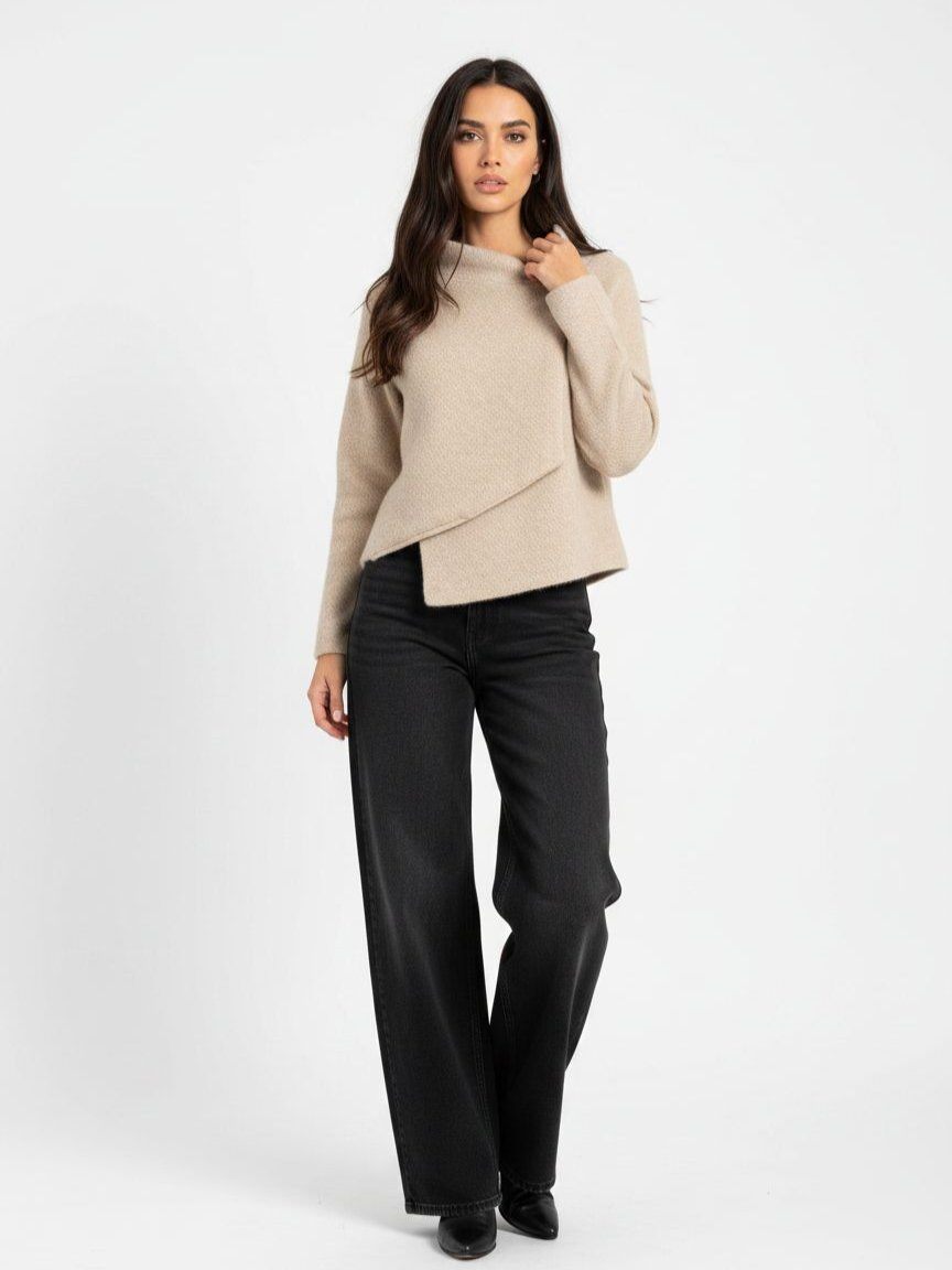 A woman modeling a light beige asymmetrical wrap-front knit sweater with black wide-leg trousers, standing against a plain white studio background.