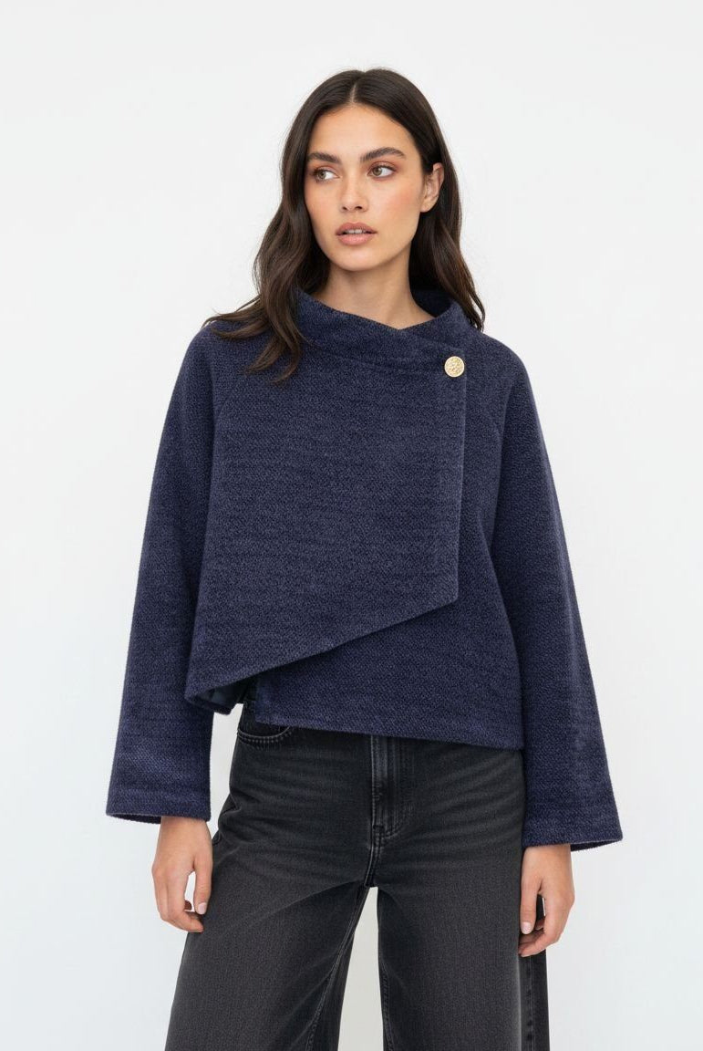 A woman modeling a navy blue textured knit wrap jacket with a single gold button closure, paired with black jeans, against a plain white studio background.