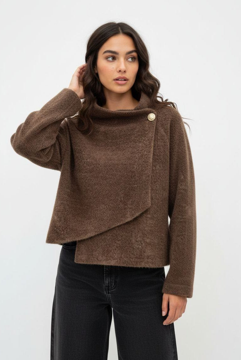 A woman modeling a brown textured knit asymmetrical wrap jacket with a single decorative gold button, paired with black wide-leg jeans, against a plain white studio background.