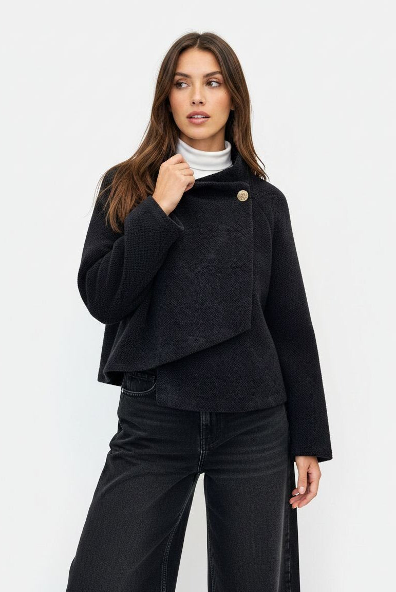 A woman modeling a black asymmetrical wrap-style jacket with a single gold-tone button, worn over a white turtleneck and black wide-leg jeans, against a plain white studio background.