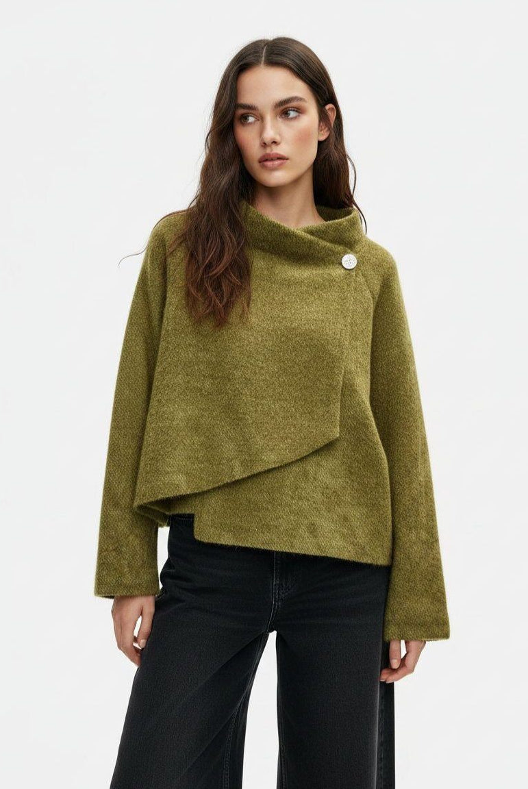 A woman wearing a textured olive green asymmetrical wrap sweater with a single decorative button, paired with black wide-leg jeans, against a plain white studio background.