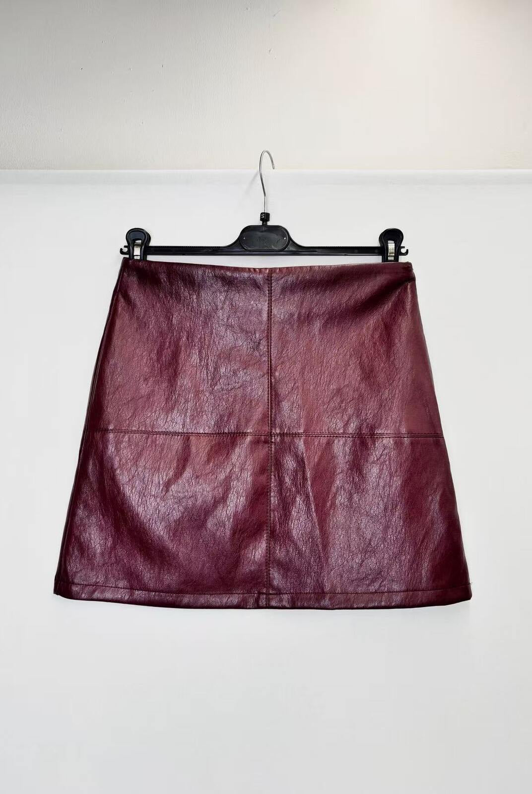 A dark burgundy faux leather A-line mini skirt hanging on a black hanger against a plain white wall.