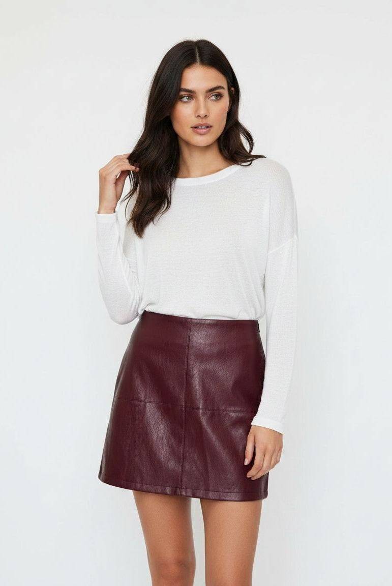 A woman modeling a white long-sleeve knit top and a dark burgundy faux-leather mini skirt against a plain white studio background.