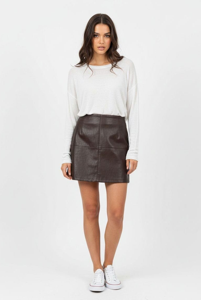 A woman modeling a dark brown faux-leather mini skirt paired with a white long-sleeve top and white sneakers, standing against a plain white studio background.