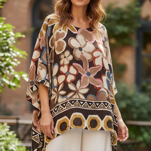A woman stands outdoors in a courtyard wearing a loose, short-sleeve patterned top with a geometric border and white wide-leg pants.