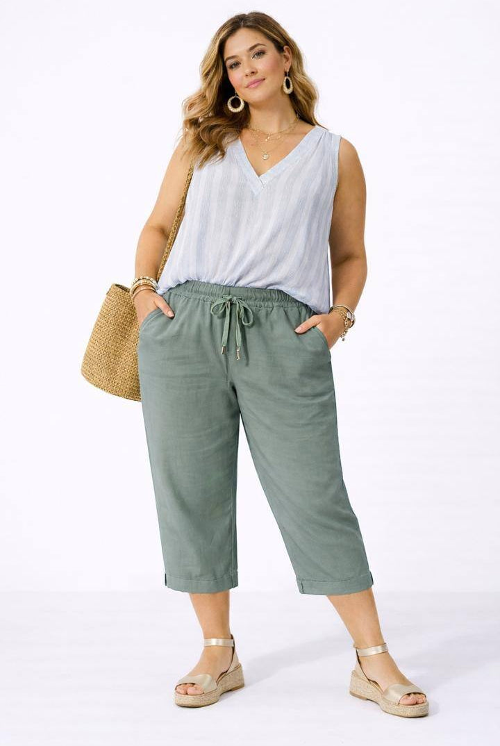 Woman wearing a white sleeveless top and olive green pants with a straw bag on a white background