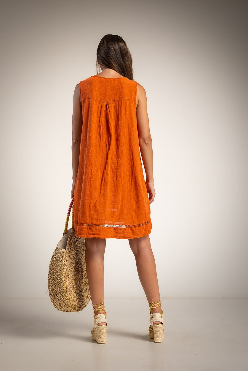 A woman is shown from behind wearing a sleeveless orange dress and holding a woven straw tote bag against a neutral studio background.