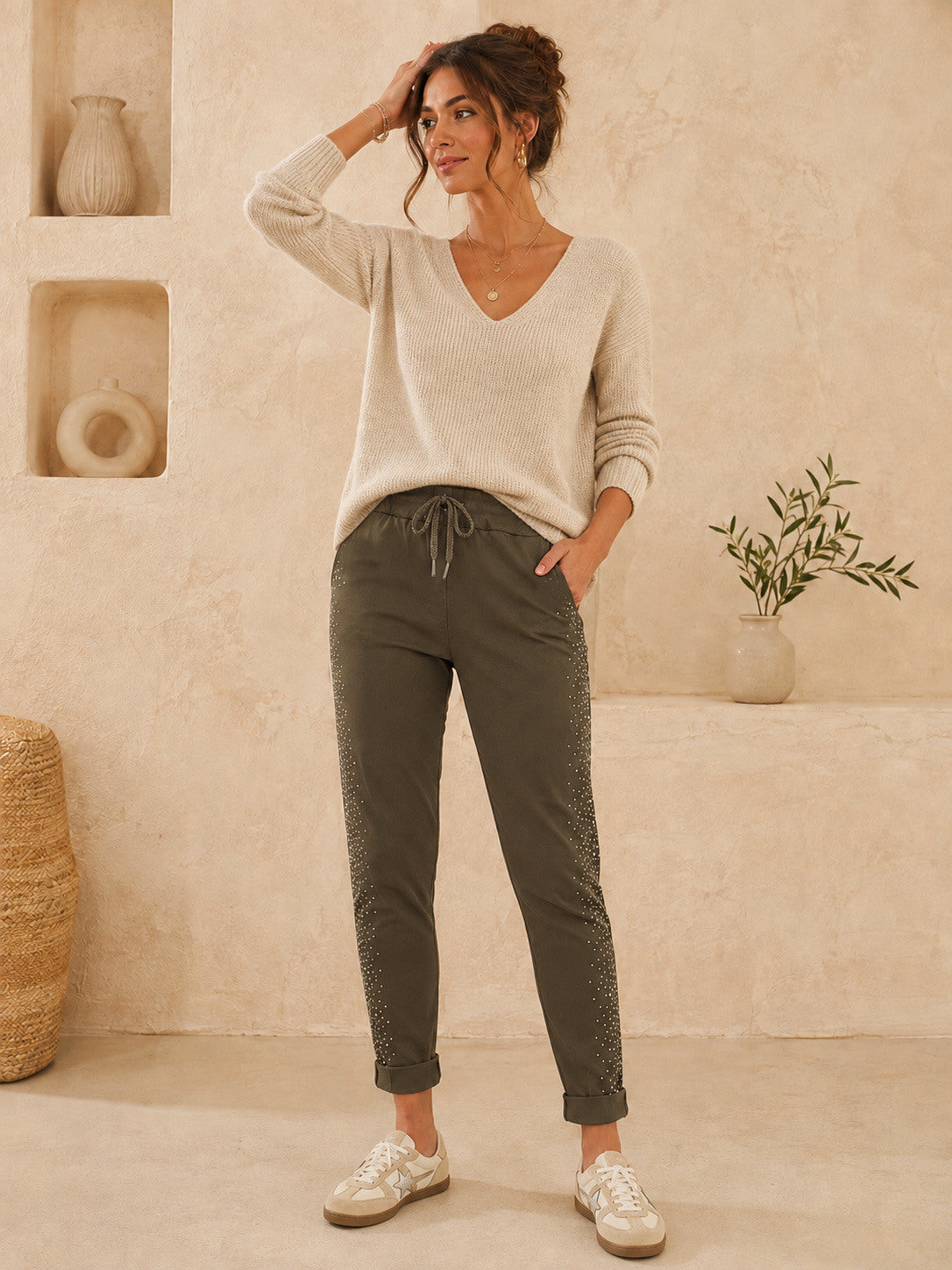 A woman stands in a warm, neutral-toned room wearing a light beige V-neck knit sweater, olive green drawstring pants with textured side panels, and white sneakers.