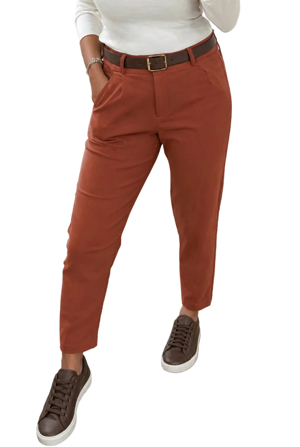 A person wearing rust-colored cropped trousers with a dark brown belt, a white long-sleeve top, and brown sneakers