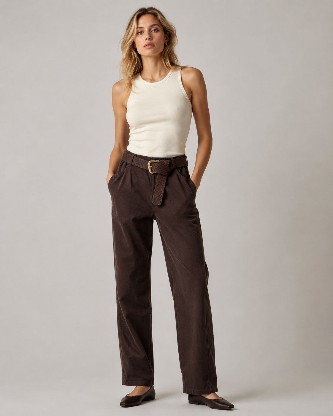 A woman modeling dark brown wide-leg trousers with a matching woven belt, paired with a cream sleeveless top and dark flats, against a neutral gray studio background.