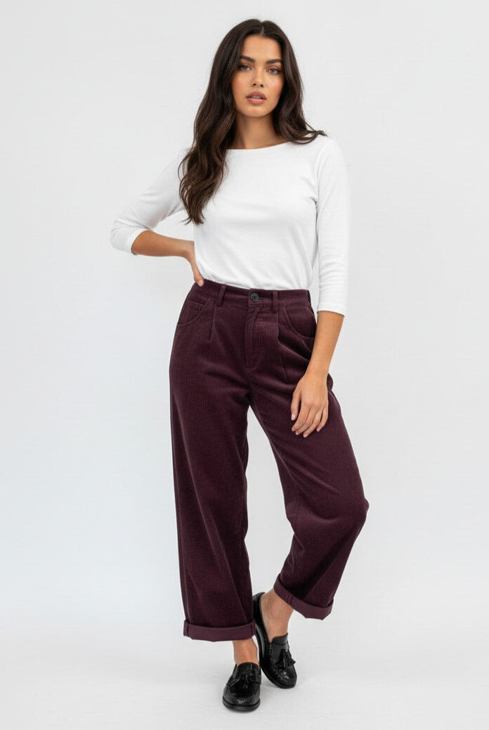 A woman modeling a pair of dark burgundy corduroy wide-leg cropped pants with a white long-sleeve top and black loafers, standing against a plain white studio background.