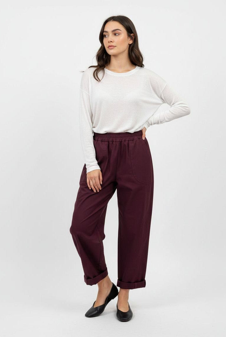 A woman modeling a white long-sleeve top and dark burgundy cropped trousers with black flats, standing against a plain light gray studio background.