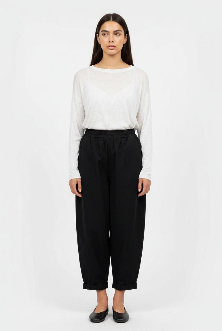 A woman stands against a plain light gray studio background wearing a white long-sleeve top and loose black cropped pants with black flats.