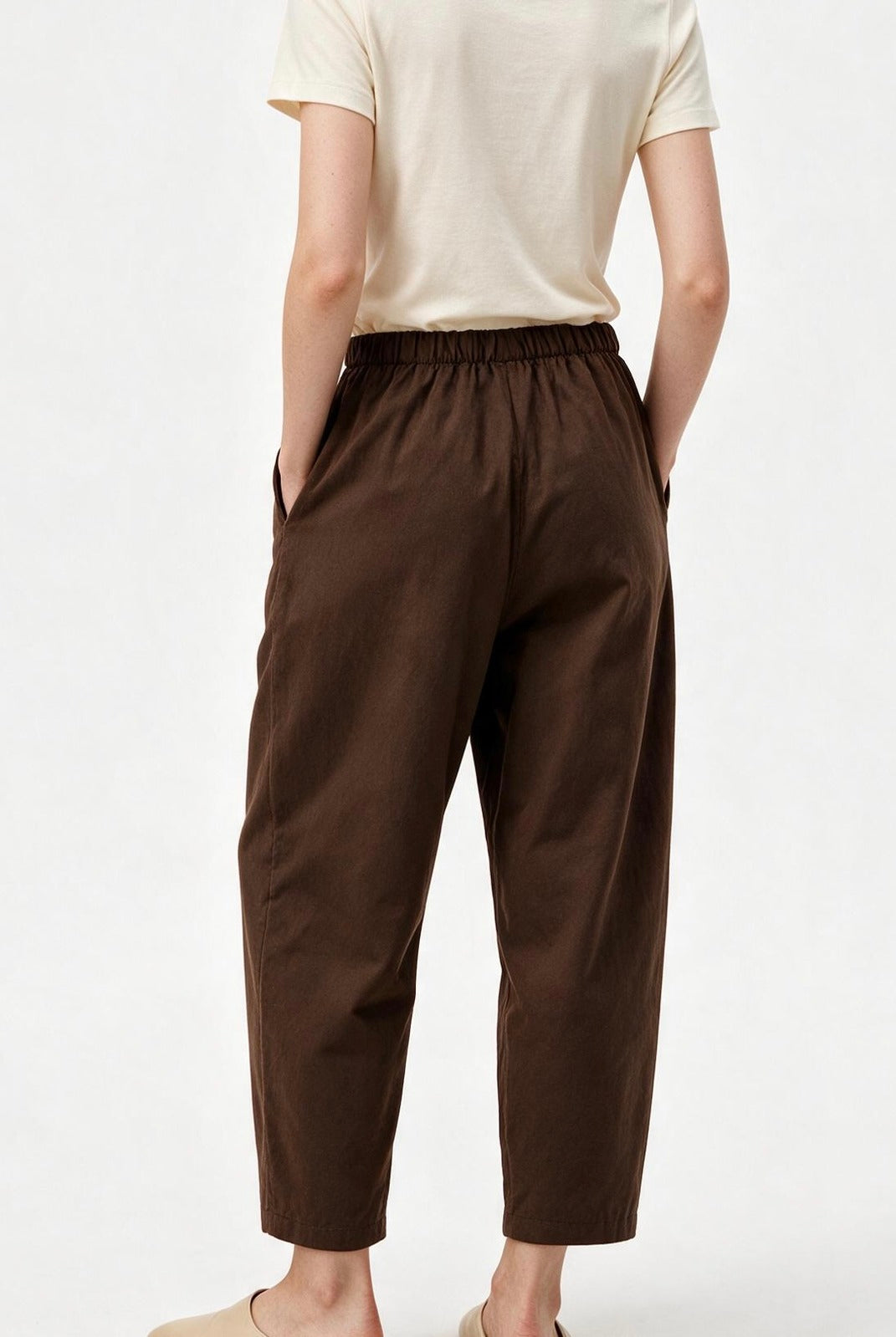A person shown from the back wearing dark brown cropped pants with an elastic waistband and side pockets, paired with a light cream short-sleeve top and beige slip-on shoes, against a plain white studio background.