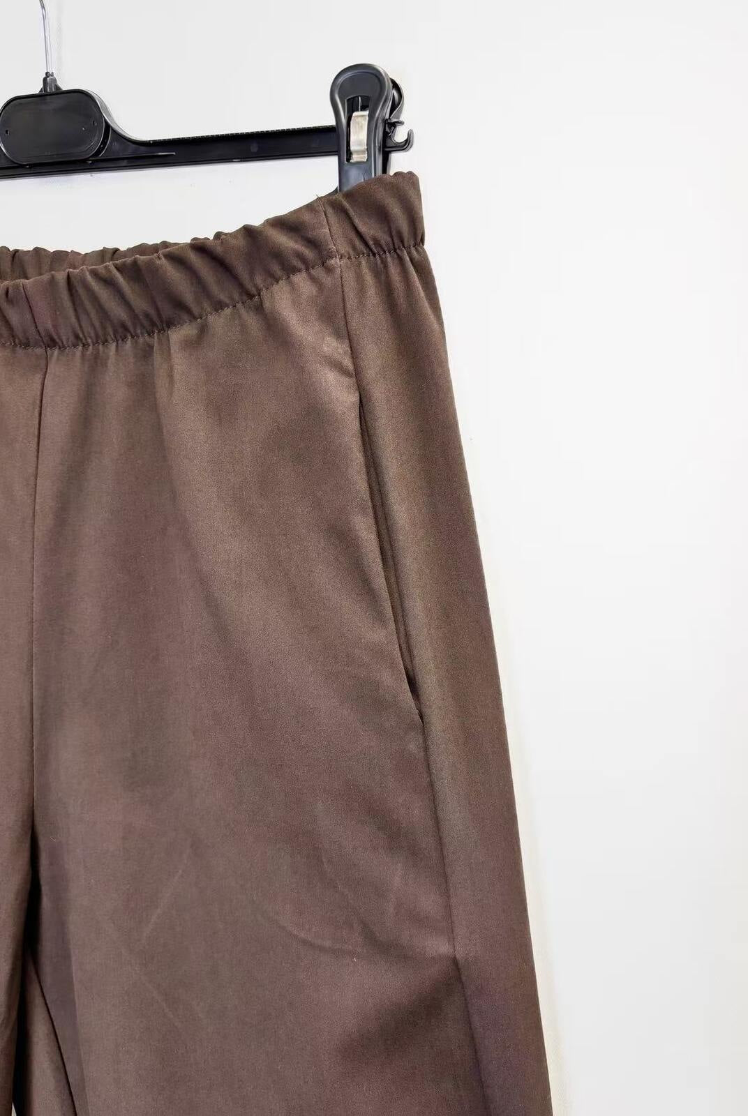 Brown wide-leg trousers with an elastic waistband, hanging on a black hanger against a plain white wall.