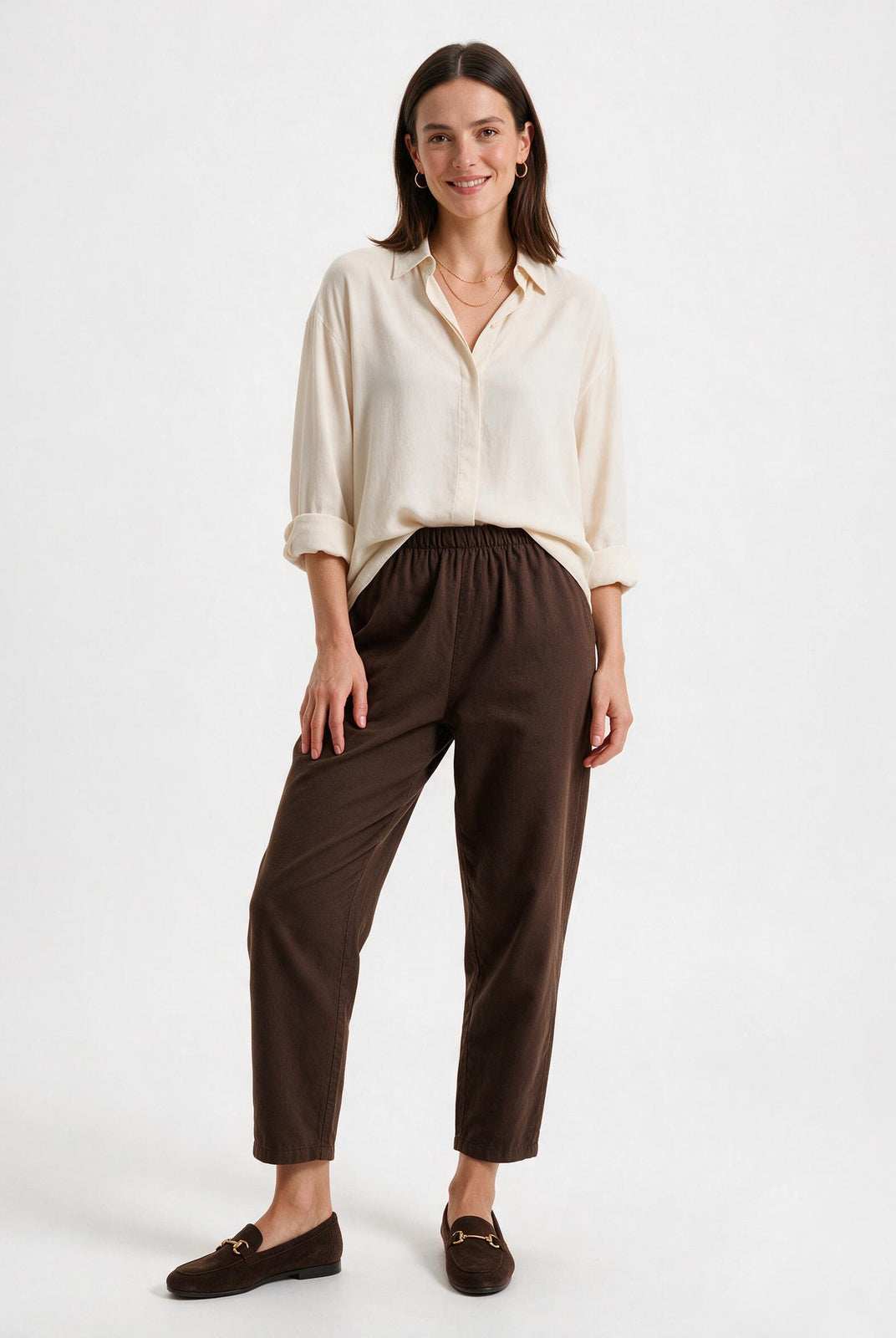 A woman modeling a cream-colored long-sleeve button-up shirt and dark brown cropped trousers with matching brown loafers, set against a plain white studio background.