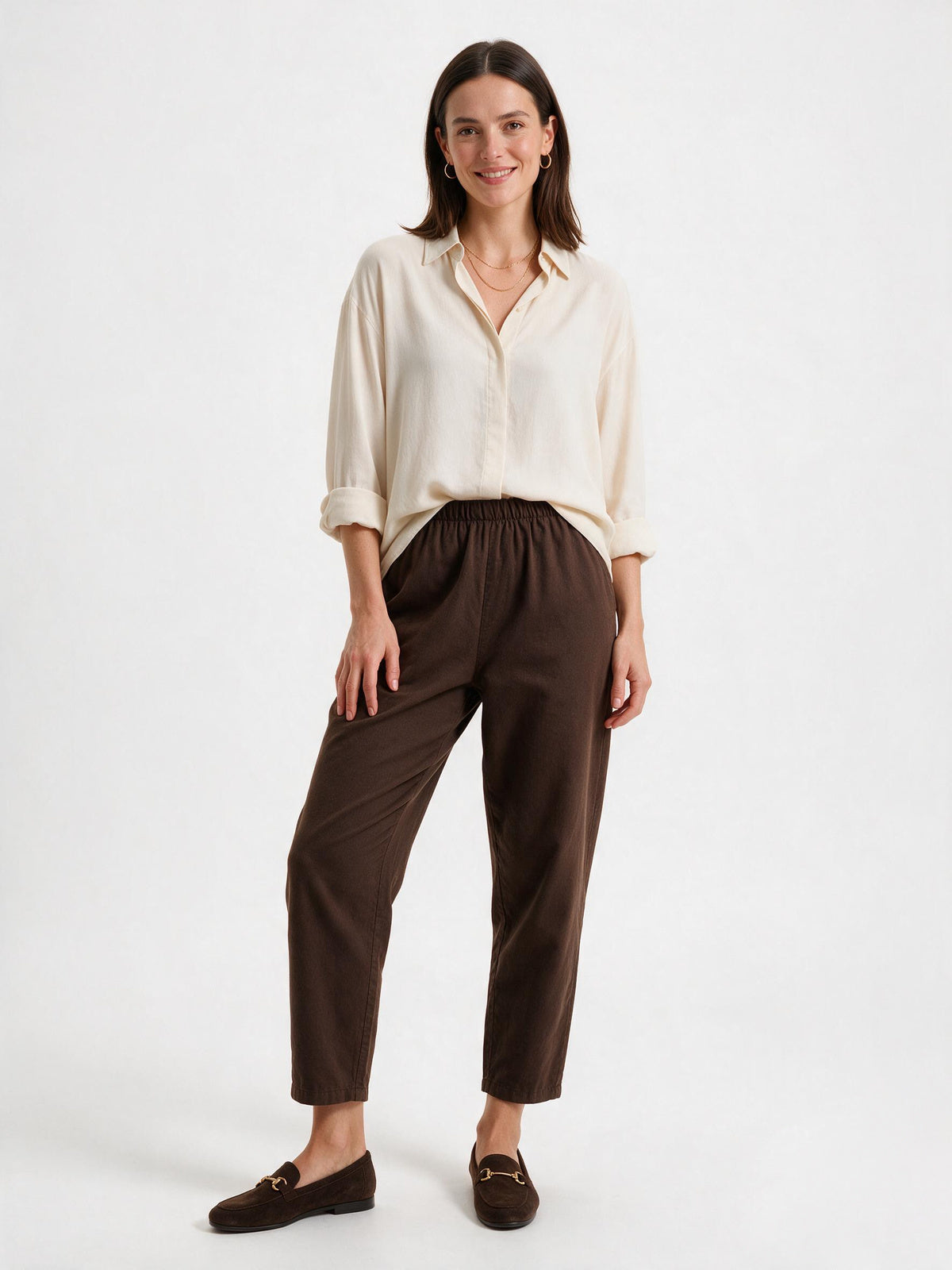 A woman modeling a cream-colored long-sleeve button-up shirt and dark brown cropped trousers with matching brown loafers, set against a plain white studio background.