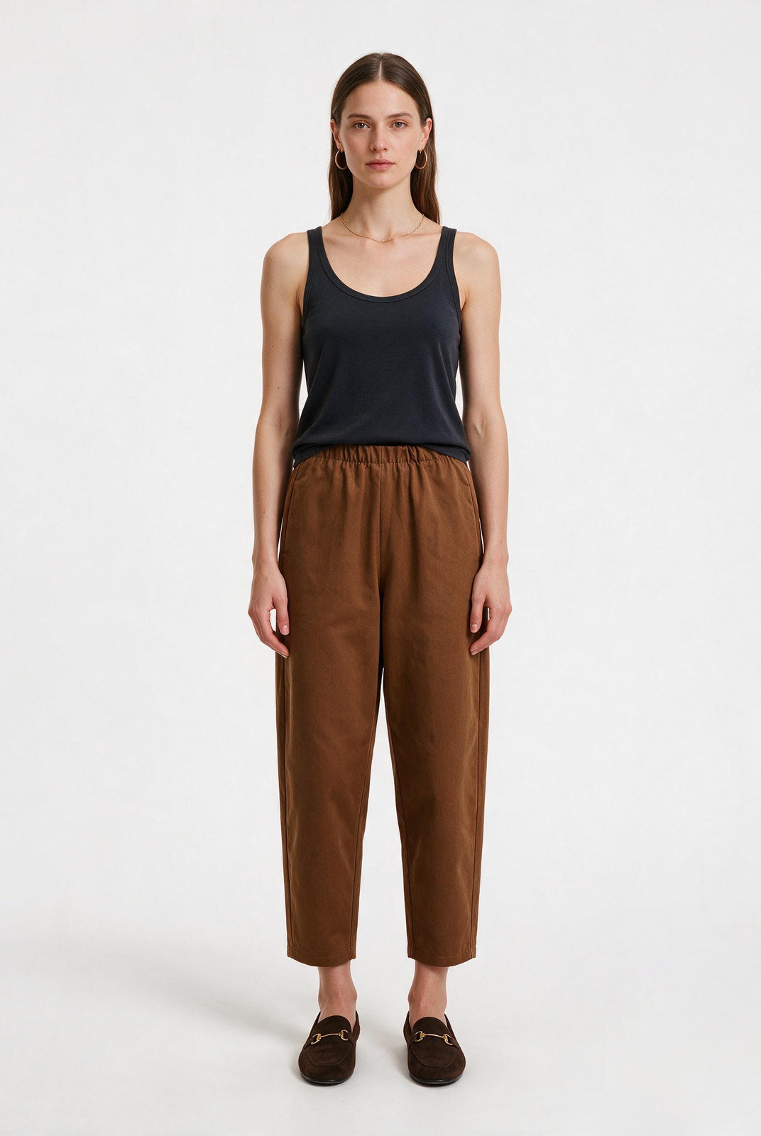 A woman stands against a plain light gray studio background wearing a dark navy sleeveless tank top, brown cropped trousers, and dark brown loafers.