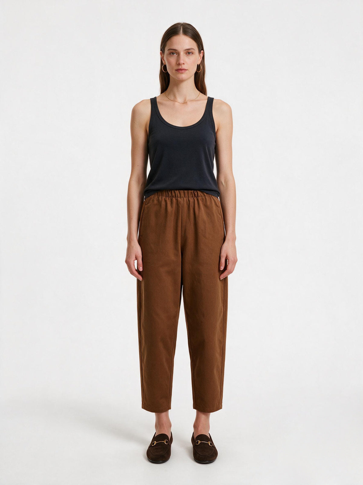 A woman stands against a plain light gray studio background wearing a dark navy sleeveless tank top, brown cropped trousers, and dark brown loafers.