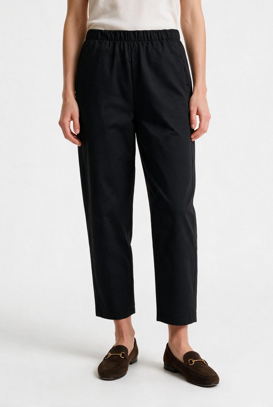 A model is shown from the waist down wearing black cropped trousers with an elastic waistband, paired with brown suede loafers and a cream top, against a plain white studio background.