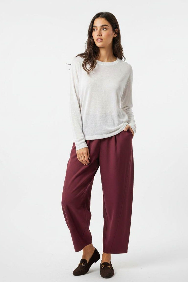 A woman modeling a white long-sleeve knit top and wide-leg burgundy cropped pants with dark loafers, photographed against a clean white studio background.