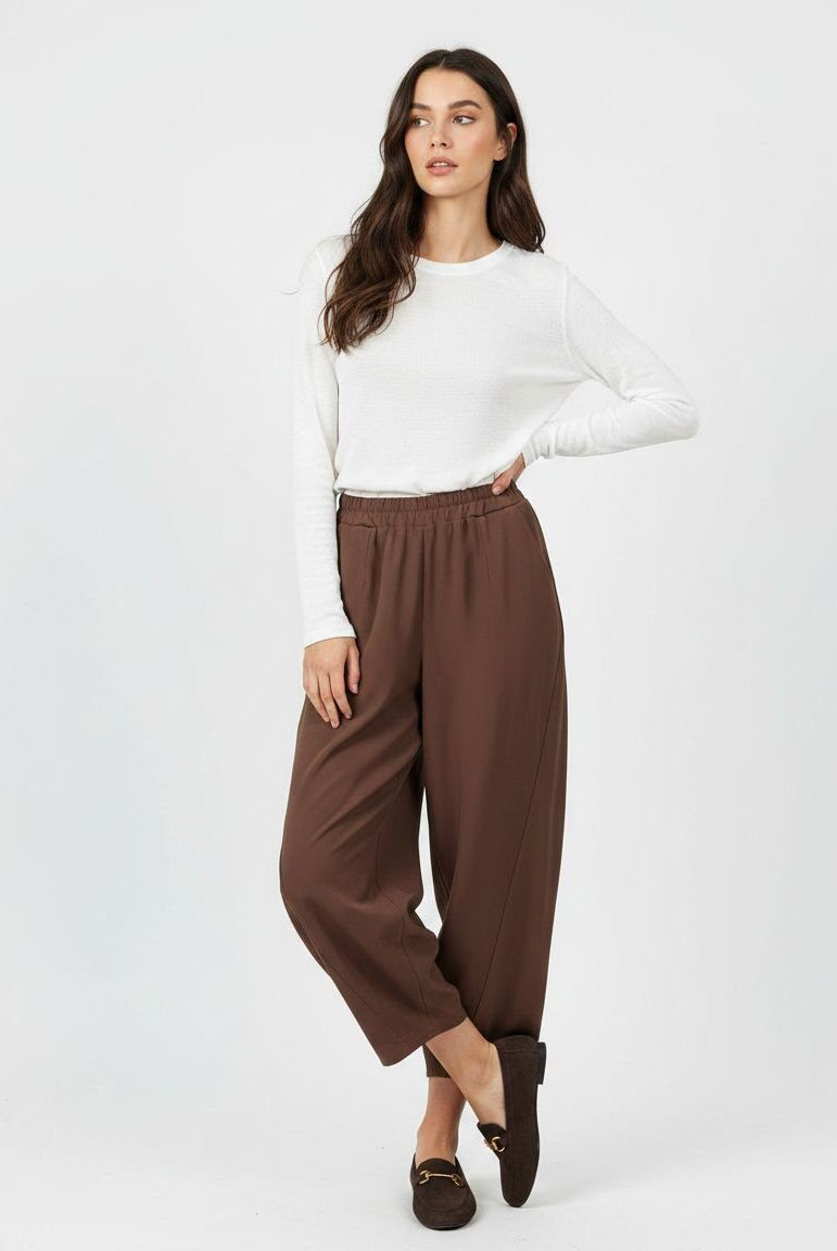 A woman modeling a white long-sleeve knit top and brown wide-leg cropped pants with dark brown loafers, standing against a light gray studio background.