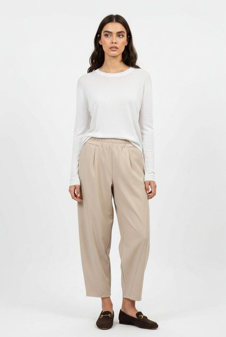 A woman stands against a plain light gray studio background wearing a white long-sleeve top, beige wide-leg cropped pants, and dark brown loafers.