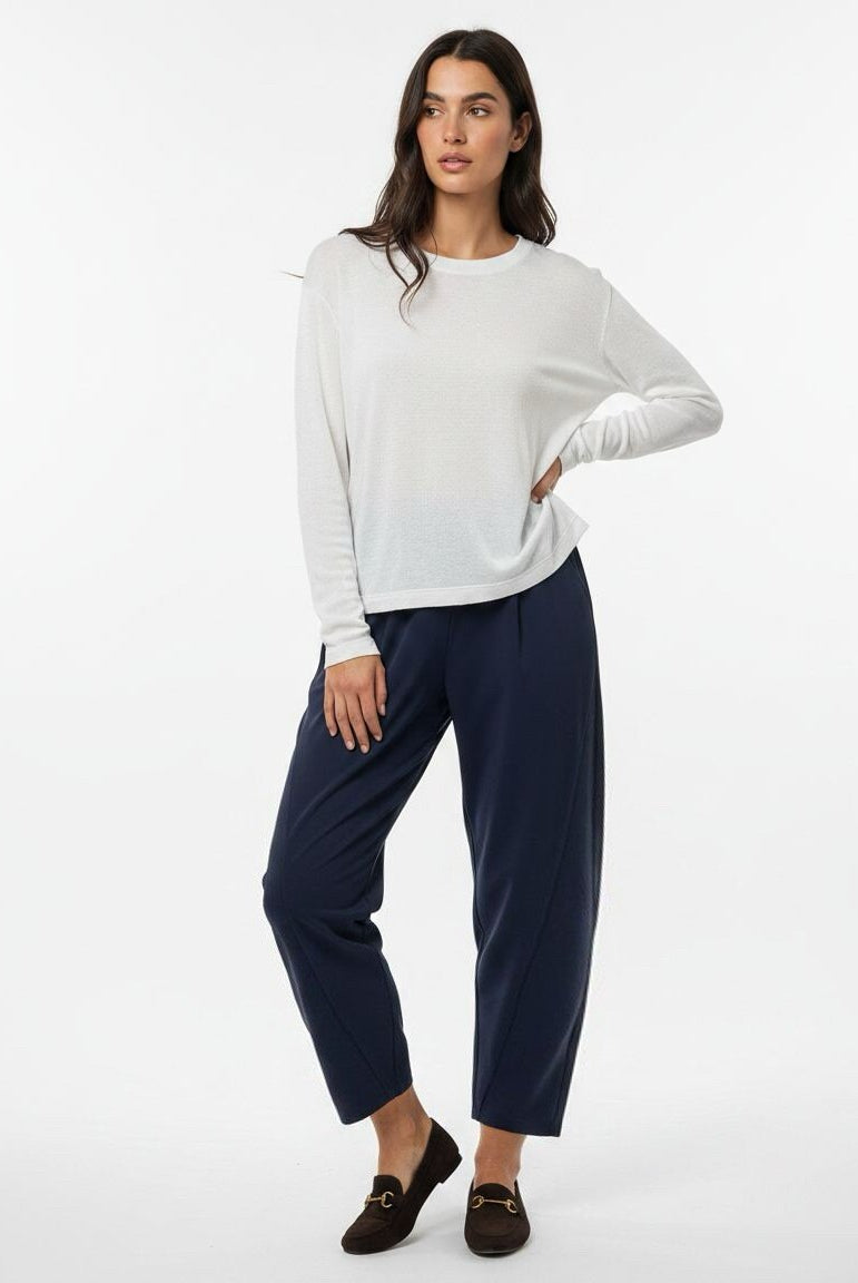 A woman modeling a white long-sleeve crewneck top and navy blue cropped trousers, paired with dark brown loafers, against a plain white studio background.