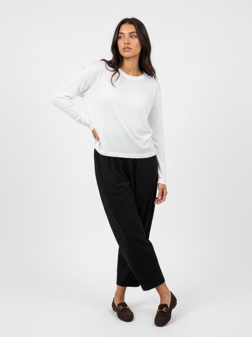 A woman modeling a white long-sleeve crewneck top and black cropped trousers, paired with dark brown loafers, against a plain white studio background.