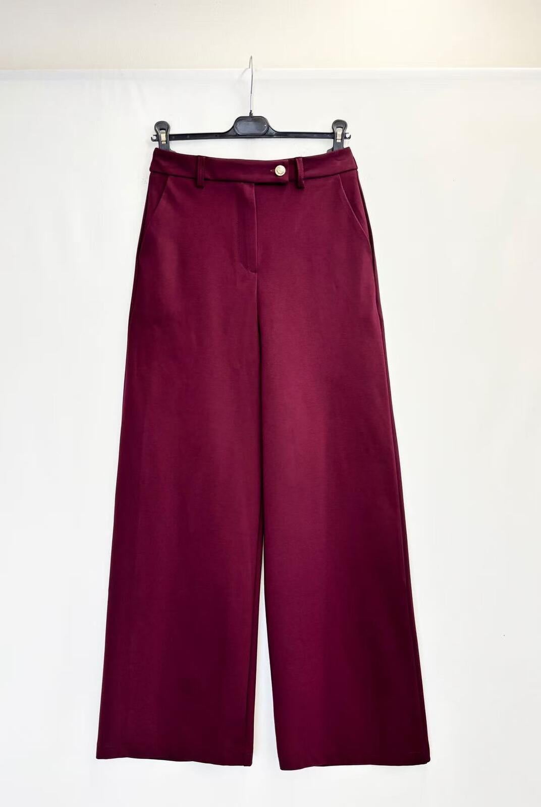 A pair of wide-leg trousers in deep burgundy, hanging on a black hanger against a plain white backdrop.