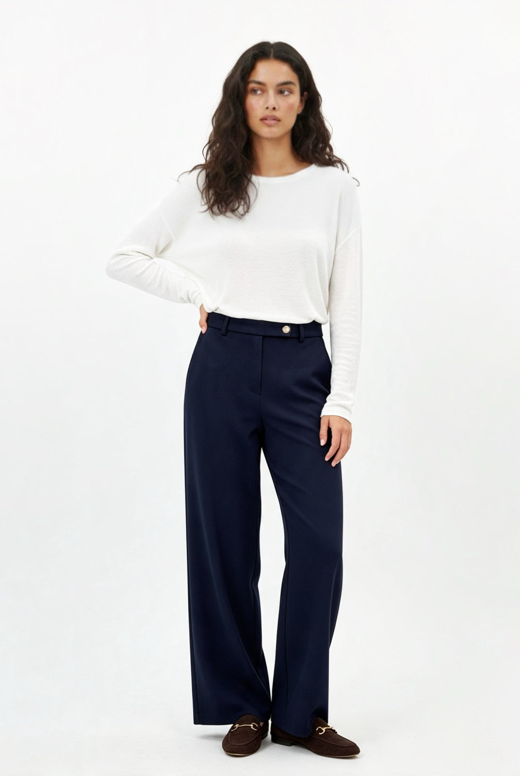 A woman modeling a white long-sleeve crewneck top and navy blue trousers, paired with dark brown loafers, against a plain white studio background.