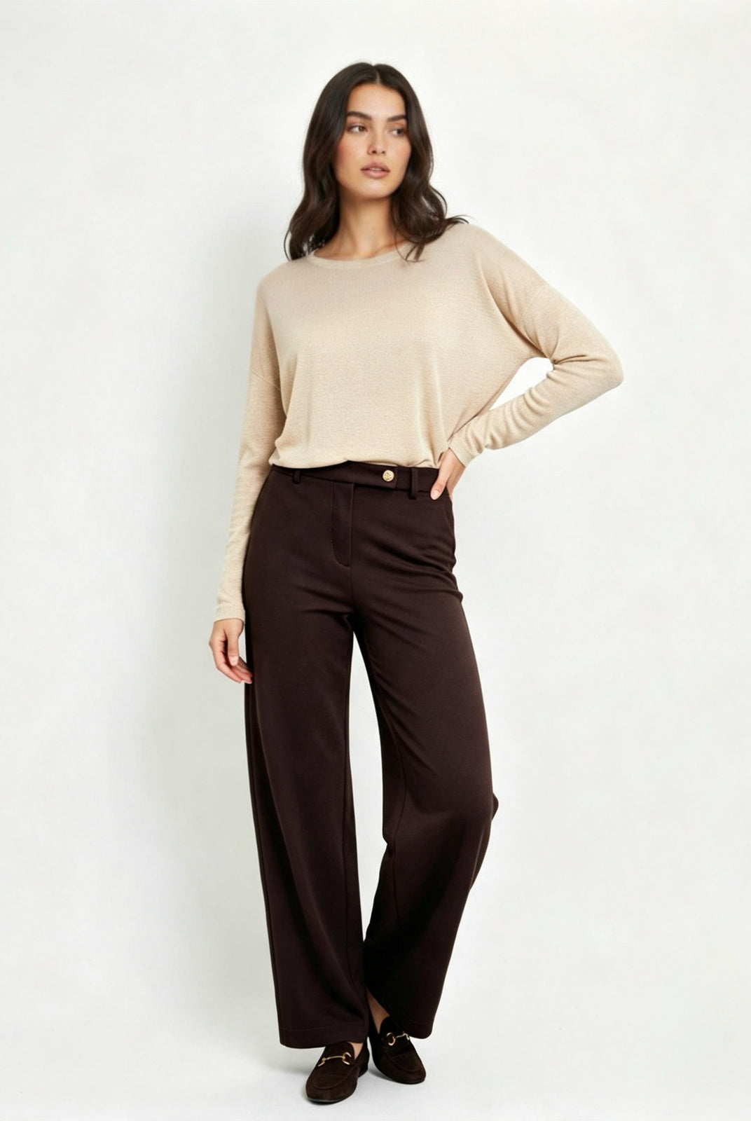 A woman modeling a beige long-sleeve knit top and dark brown wide-leg trousers against a plain light gray studio background.