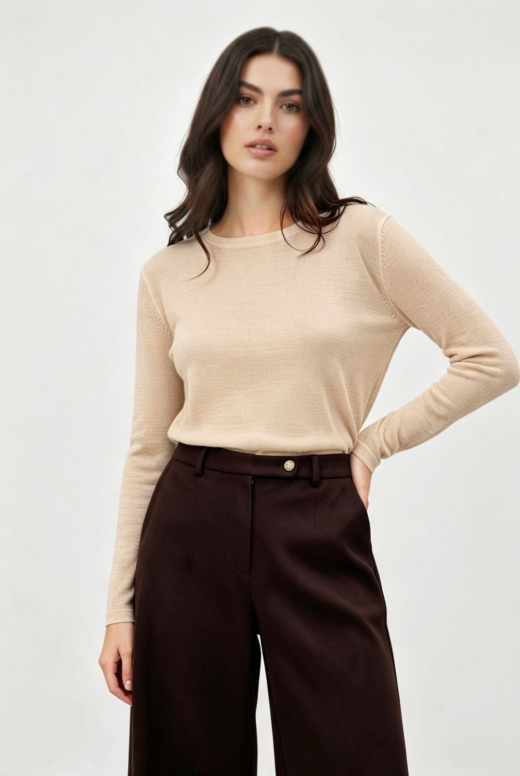 A woman modeling a beige long-sleeve crewneck knit top paired with dark brown wide-leg trousers, photographed against a clean light gray studio background.